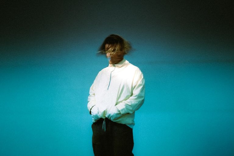 Australian electronic band Crooked Colours member wearing a white jacket, standing against a teal background in a press photo.
